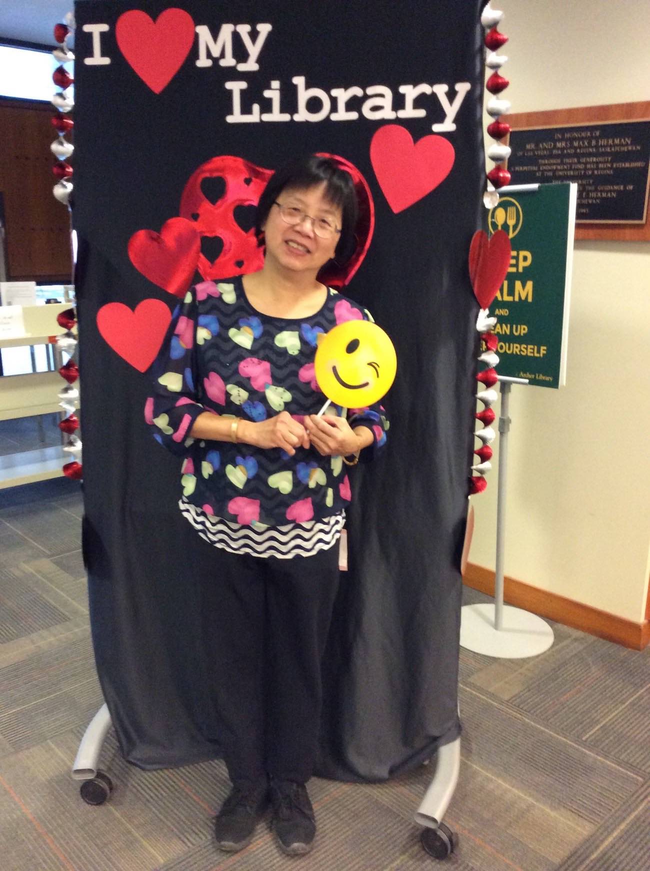 Archives staff member Kieu Ma stands in front of the I love my Library banner holding up a winking smiley face emoji on a stick and smiling
